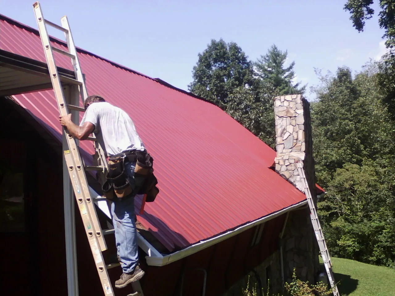 Roofer on a ladder beside a red metal roof and stone chimney — Soffit & Fascia Repair by Roofing Brothers in Abercrombie